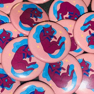 May include: A collection of pink button pins featuring a stylized illustration of a purple cat with blue swirls and white stars.