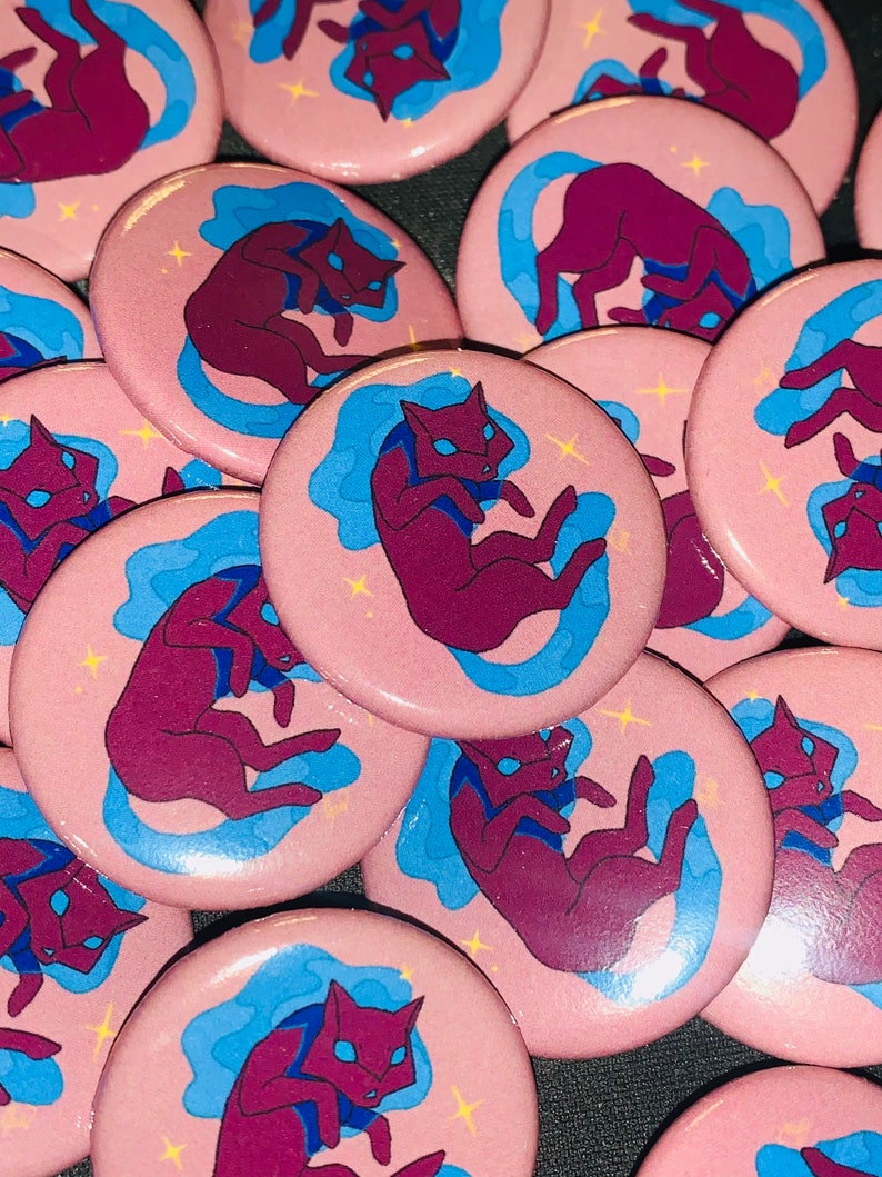 May include: A collection of pink button pins featuring a stylized illustration of a purple cat with blue swirls and white stars.