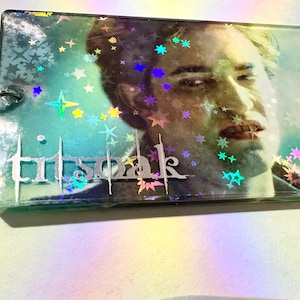 Puede incluir: Un llavero de plástico transparente con una imagen holográfica de un hombre con una inscripción plateada que dice "tristoak" y un fondo de estrellas.