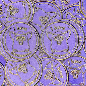 Pode incluir: Um close-up de vários patches de tecido roxo com bordados dourados. Cada patch apresenta um personagem de desenho animado com olhos grandes e o texto "Eu tenho" seguido de "depressão" ou "ansiedade".
