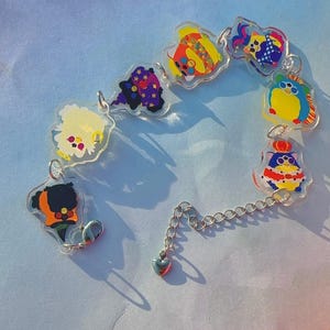 Könnte beinhalten: Ein farbenfrohes Charm-Armband mit Cartoon-Charakter-Designs. Das Armband hat eine silberne Kette mit einem herzförmigen Anhänger am Ende. Die Charms sind in verschiedenen Farben, darunter Gelb, Blau, Lila und Orange.