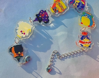 Puede incluir: Una colorida pulsera de dijes con diseños de personajes de dibujos animados. La pulsera tiene una cadena plateada con un dije en forma de corazón al final. Los dijes son de varios colores, incluyendo amarillo, azul, morado y naranja.