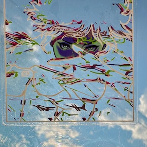 May include: Square sticker with an illustration of a person's face with green eyes and purple skin. The artwork has a blue background with white, red, and green abstract lines. The sticker is in a clear plastic case.