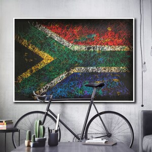 May include: A framed artwork featuring the South African flag, rendered with colorful scribbled lines on a black background. The artwork is displayed above a black bicycle and a table with decorative items, set against a gray wall.