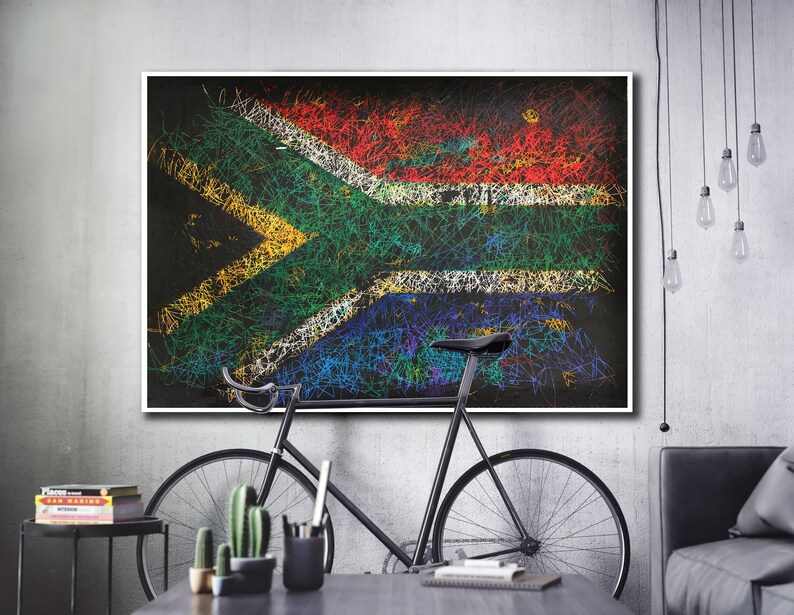 May include: A framed artwork featuring the South African flag, rendered with colorful scribbled lines on a black background. The artwork is displayed above a black bicycle and a table with decorative items, set against a gray wall.