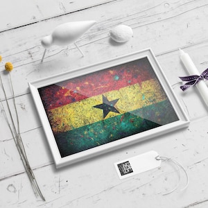 Puede incluir: Una impresi&oacute;n enmarcada de la bandera de Ghana, con un aspecto desgastado y texturizado. La bandera es roja, amarilla y verde con una estrella negra en el centro. El marco es blanco y la impresi&oacute;n est&aacute; sobre una superficie de madera blanca.