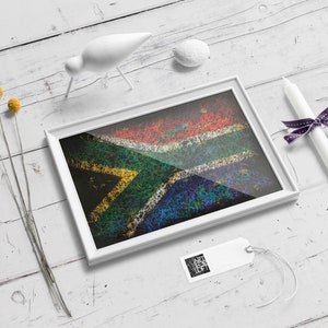 May include: A framed artwork featuring the South African flag, rendered with a textured, colorful design. The white frame sits on a white wooden surface, accompanied by decorative elements like a white bird figurine, a candle, and a tag.