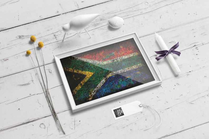 May include: A framed artwork featuring the South African flag, rendered with a textured, colorful design. The white frame sits on a white wooden surface, accompanied by decorative elements like a white bird figurine, a candle, and a tag.