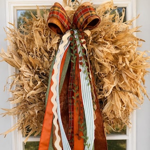 May include: A circular wreath constructed from dried corn husks, featuring a large decorative bow at the top. The bow consists of multiple ribbons in orange, green, and plaid patterns, with long, trailing streamers in various textures and colors.