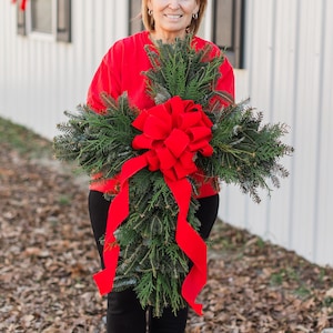 May include: A woman in a red sweater holds a large green Christmas cross with a red velvet bow. The cross is made of evergreen branches and is decorated with a large red bow.
