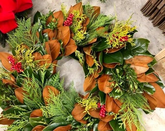 Könnte beinhalten: Ein Weihnachtskranz aus Magnolienblättern, Zedern und roten Beeren. Der Kranz ist in einem natürlichen, rustikalen Stil gehalten und wäre eine schöne Ergänzung für jede Weihnachtsdekoration.