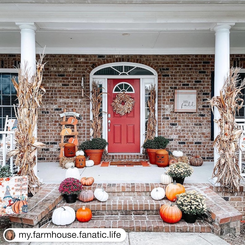 May include: A red front door with a fall wreath and a sign that says "hello fall" on a brick house. The porch is decorated with pumpkins, gourds, and fall foliage.