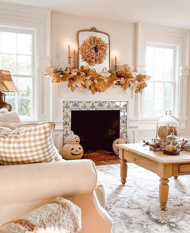 May include: A white fireplace mantel decorated with a fall-themed garland of brown leaves and a straw wreath. The fireplace has a black interior and is tiled with blue and white patterned tiles. Two carved pumpkin figurines are in front of the fireplace. A wooden coffee table with a glass cloche containing a small pumpkin sits in front of the fireplace.