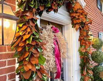 May include: A lush magnolia leaf garland featuring dual-toned green and copper-brown foliage draped over a white door frame. A textured, light-colored wreath with a long, vibrant magenta ribbon hangs on the center of the wooden door.
