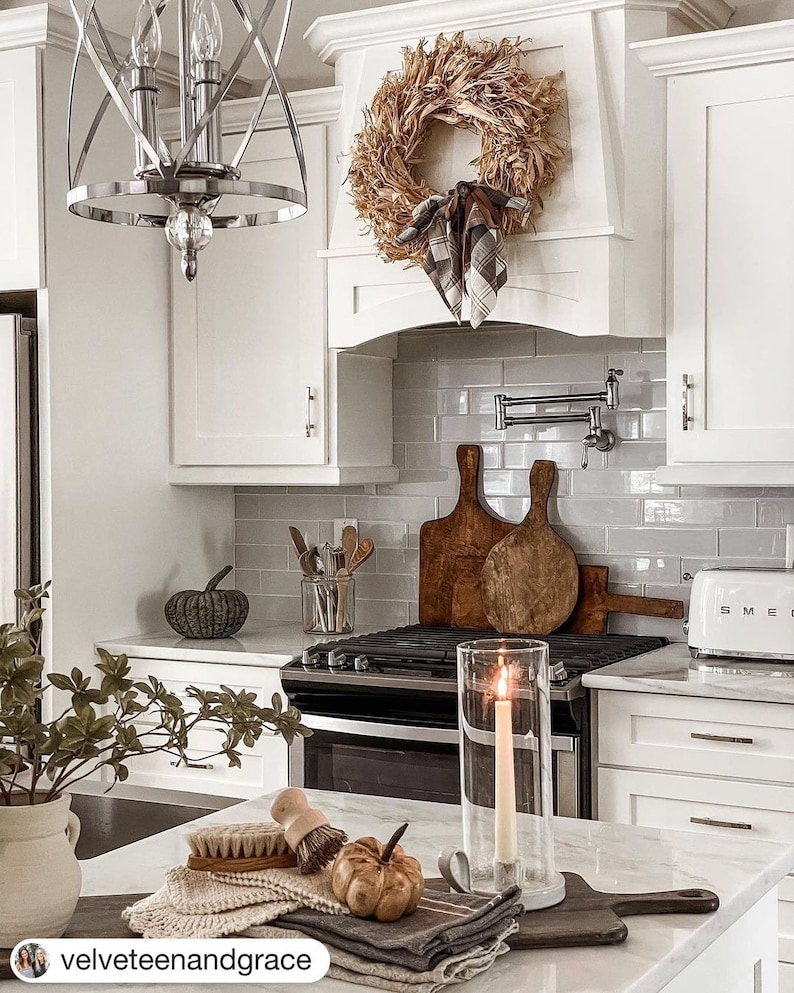 May include: A white kitchen with white cabinets, a white tiled backsplash, and a stainless steel stove. There is a wreath made of dried corn husks hanging above the stove. There is a small pumpkin on the counter and a candle in a glass holder.