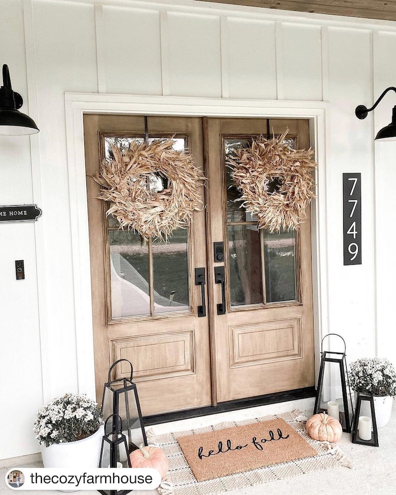 May include: A white farmhouse exterior with a double wooden door decorated with two large, dried corn husk wreaths. The door has a black door handle and a welcome mat that reads "hello fall". There are two black lanterns on the porch, one with a pumpkin inside. The house number is 7749.