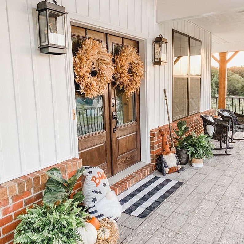 May include: A white farmhouse with a wooden front door decorated with two large straw wreaths. The door has glass panels and a black door handle. There is a black and white striped doormat on the porch. There are two black wicker chairs on the porch with white cushions. There is a potted plant on the porch and a gnome with a "Boo" sign.
