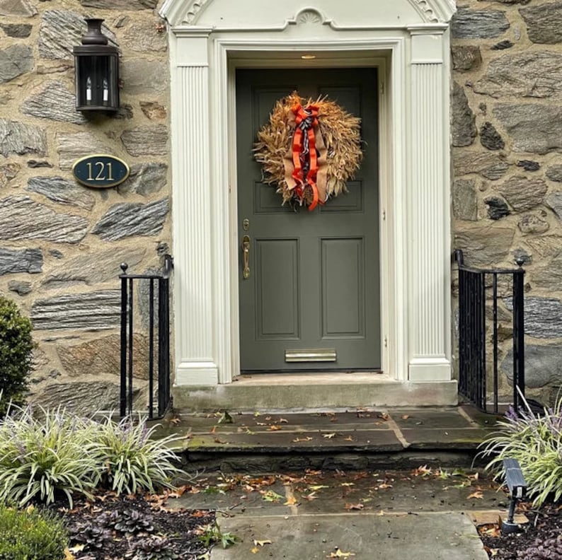 May include: A green front door with a brown and orange wreath. The door has a brass door handle and a brass door knocker. The house number 121 is on the stone wall to the left of the door.