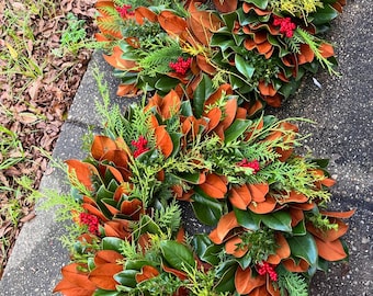 Könnte beinhalten: Drei Kränze aus Magnolienblättern, Zedern und roten Beeren. Die Kränze sind übereinander gestapelt, der größte unten und der kleinste oben.