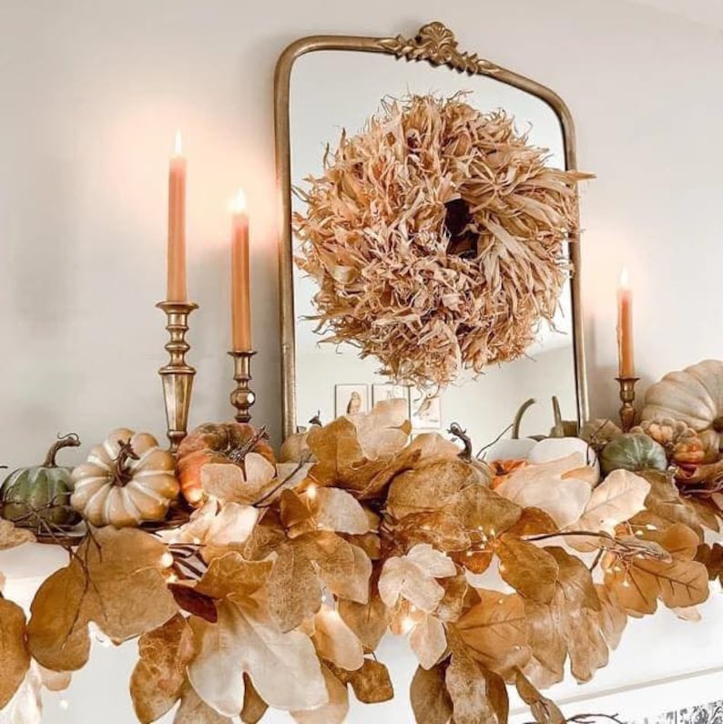 May include: A gold-framed mirror with a dried corn husk wreath reflected in it. The mantel is decorated with a garland of brown leaves, white pumpkins, and small lights.