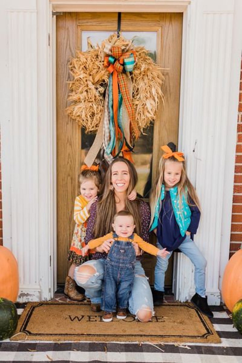 May include: A family of four poses in front of a door with a fall-themed wreath. The mother is wearing a blue denim shirt and jeans. The children are wearing a yellow shirt and jeans, a blue shirt and jeans, and a blue shirt and overalls. The doormat says "Welcome".