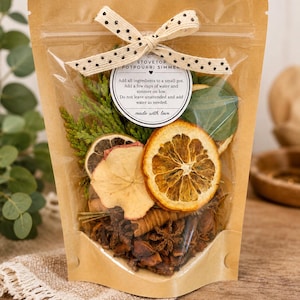 May include: A clear-fronted kraft paper pouch containing stovetop potpourri ingredients including dried orange slices, apple slices, star anise, cinnamon sticks, and greenery. A cream ribbon with black dots ties the top, and a circular label reads: STOVETOP POTPOURRI SIMMER. Add all ingredients to a small pot. Add a few cups of water and simmer on low. Do not leave unattended and add water as needed. made with love.