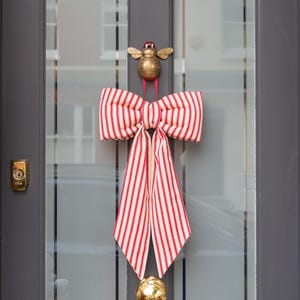 May include: A door knocker with a gold bee design, a red and white striped bow, and a gold sphere. The door is dark grey with glass panels. The bow is a decorative element.