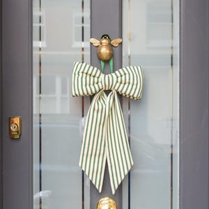 May include: A decorative door bow with a green and white striped pattern, hanging on a grey door. The bow is adorned with a gold-coloured bee and a matching gold door knob.