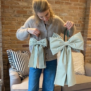 May include: Two large decorative bows with green and white stripes. The bows are held up by a person. A striped cushion and a beige sofa are in the background. The bows are made of fabric and have long, flowing tails.