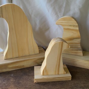 May include: Three wooden wave-shaped sculptures, each mounted on a rectangular base. The sculptures are made of light-colored wood with visible grain patterns. The largest wave is on the left, with two smaller ones to the right.