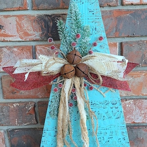 May include: A wooden Christmas tree decoration with a rusty star topper. The turquoise tree features musical notes and is adorned with faux greenery, red berries, and rustic bells. A red and cream ribbon adds a festive touch.