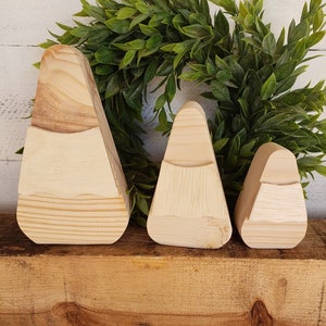 May include: Three wooden candy corn ornaments in different sizes. The ornaments are made of natural wood and have a light brown color. The ornaments are arranged in a row on a wooden surface.