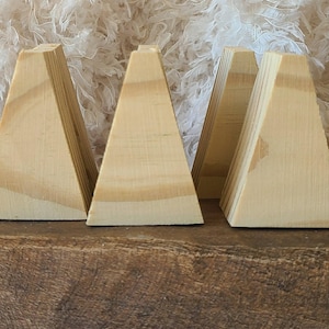 May include: A set of four light-colored wooden blocks, each shaped like a trapezoid with a flat base and a narrow top. The natural wood grain is visible on the smooth surfaces of these geometric pieces, which are arranged in a row.
