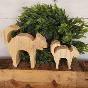 May include: Two wooden cat figurines, one large and one small, stand side-by-side on a wooden surface. The cats are carved from light-colored wood and have a simple, stylized design. A green leafy wreath is in the background.