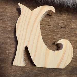 May include: A wooden cutout in the shape of a witch's shoe, crafted from light-colored wood. The shoe features a pointed toe and a curled heel, with visible wood grain. The cutout is designed for crafting or decoration.