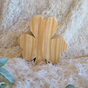 May include: A wooden shamrock, a symbol of St. Patrick's Day, is displayed. The shamrock is made of light-colored wood, with visible grain, and is set against a fluffy, white background. The clover has three heart-shaped leaves.