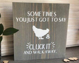 Puede incluir: Un letrero de madera gris con texto blanco que dice "Sometimes you just got to say Cluck It and walk away." Una silueta blanca de una gallina está representada en el letrero.