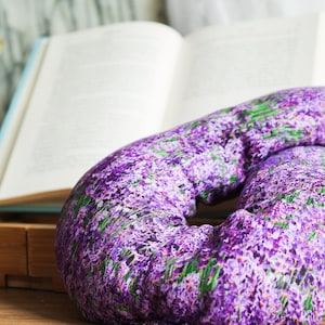 May include: A purple and green lavender-patterned neck pillow. The pillow is shaped like a donut and is lying on a wooden surface.