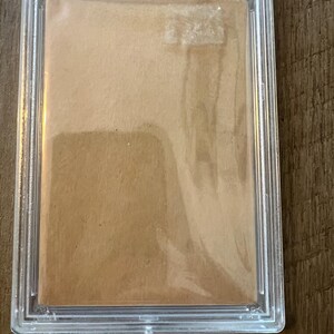 May include: A clear plastic case with a brown card inside. The case has the text "Sports Authenticator" and "Collectors Universe NASDAQ: CLCT" printed on it.