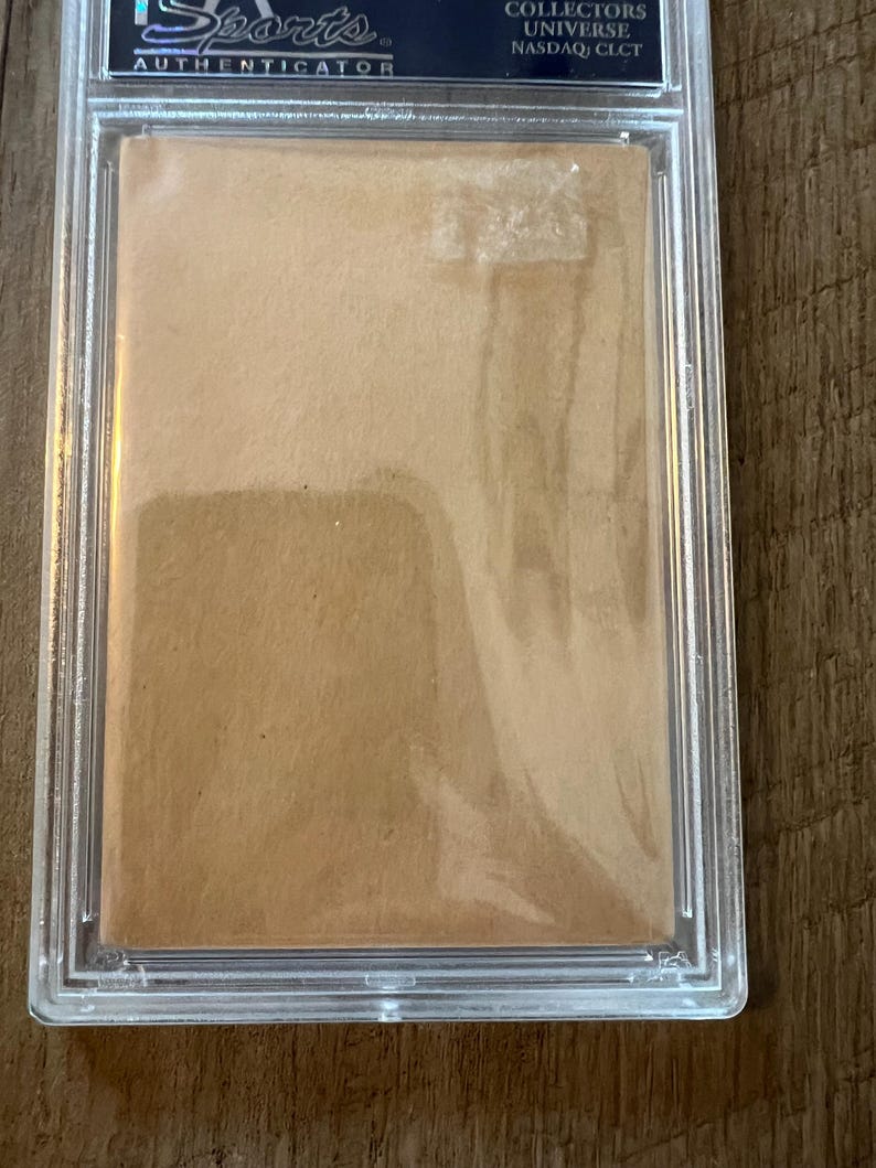 May include: A clear plastic case with a brown card inside. The case has the text "Sports Authenticator" and "Collectors Universe NASDAQ: CLCT" printed on it.