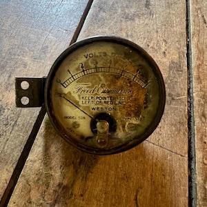 Puede incluir: Un voltímetro Freed-Eisemann Weston antiguo, modelo 506, con una carcasa de metal negro y un indicador circular. El indicador muestra "VOLTS D.C." y una aguja que indica el nivel de voltaje. El texto "KEEP POINTER TO LEFT OF RED LINE" es visible. El dispositivo está sobre una superficie de madera.