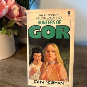 May include: A vintage paperback book titled "Hunters of Gor" by John Norman. The cover features a woman with long blonde hair and a man with dark hair. The book is displayed on a small wooden easel.
