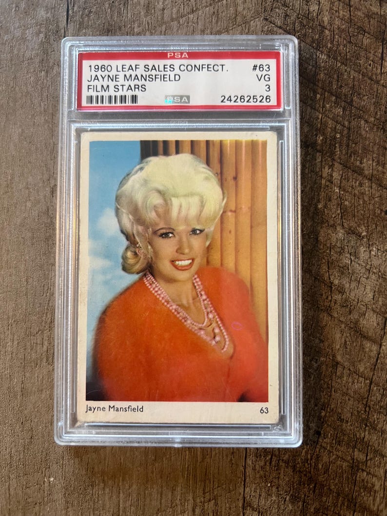 May include: A vintage trading card featuring a color photo of Jayne Mansfield, a blonde actress wearing a red sweater and a pearl necklace. The card is graded VG 3 by PSA and has the number 63 printed on it. The card is from the 1960 Leaf Sales Confection Film Stars series.