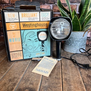 May include: A vintage Westinghouse movie light in black with a sealed beam. The light is mounted on a tripod and has a cord. The box for the light is also pictured and features a woman using a movie camera. The box reads "Westinghouse Sealed Beam Type Movie Lamp".