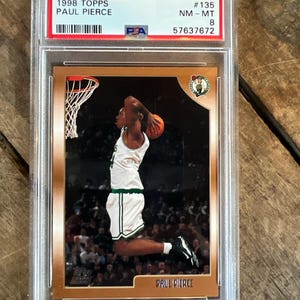 May include: A 1998 Topps Paul Pierce basketball card in a protective case. The card features a player in a white and green uniform dunking a basketball. The card is graded and labeled with the number 135.