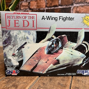 Puede incluir: Caja de un kit de modelo del caza A-Wing de Star Wars. La caja presenta el texto "Star Wars Return of the Jedi" y una ilustración del caza A-Wing en el espacio. También tiene el texto "It's a Snap" y "Golden Opportunity Kit."
