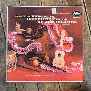 May include: A vintage vinyl record cover titled Hawaii Calls: Favorite Instrumentals of the Islands, featuring a cello, a small ukulele, a drum, and maracas adorned with a pink floral lei against a deep red background with Capitol Records logo.