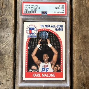 Pu&ograve; includere: Una carta da collezione NBA Hoops del 1989 con Karl Malone, un giocatore di basket degli Utah Jazz, che tiene in alto il trofeo dell'NBA All-Star Game del 1989. La carta &egrave; classificata NM-MT 8 da PSA e ha il numero #116.