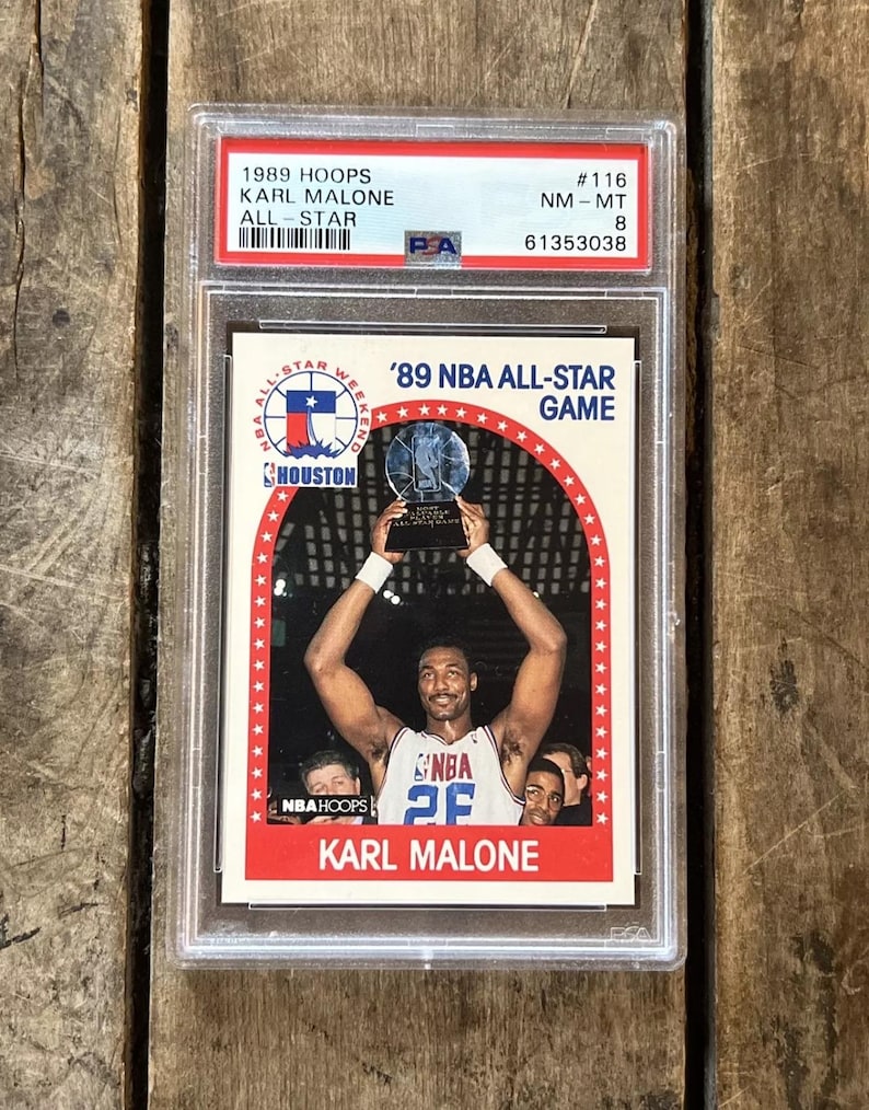 Pu&ograve; includere: Una carta da collezione NBA Hoops del 1989 con Karl Malone, un giocatore di basket degli Utah Jazz, che tiene in alto il trofeo dell'NBA All-Star Game del 1989. La carta &egrave; classificata NM-MT 8 da PSA e ha il numero #116.