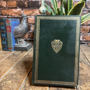 Pode incluir: Um livro verde de capa dura com uma borda dourada e um brasão dourado com o texto "VERI TAS" na capa.